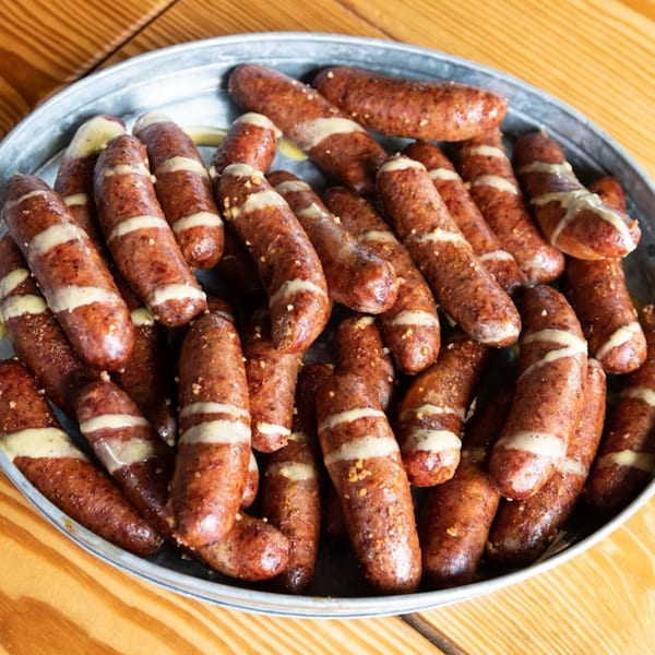 A metal tray filled with numerous cooked sausages, drizzled with a light-colored sauce, on a wooden table.
