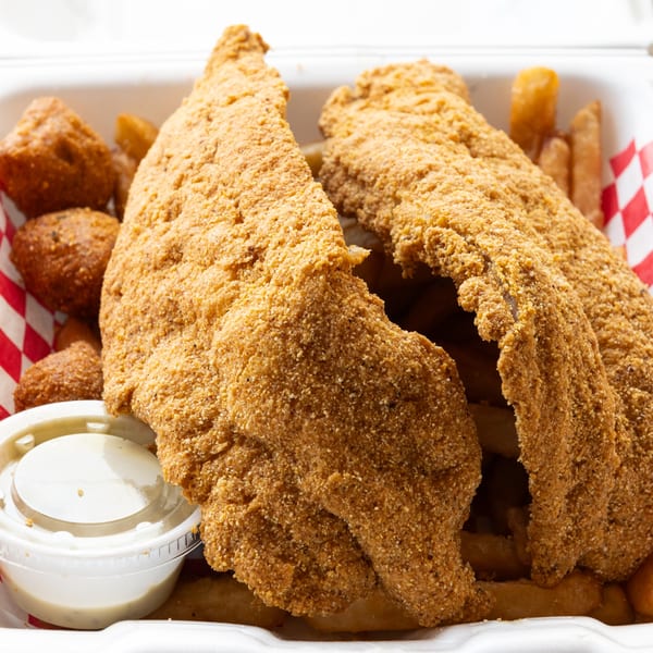 Crispy fried fish fillets, hush puppies, and french fries in a take-out container with dipping sauce.