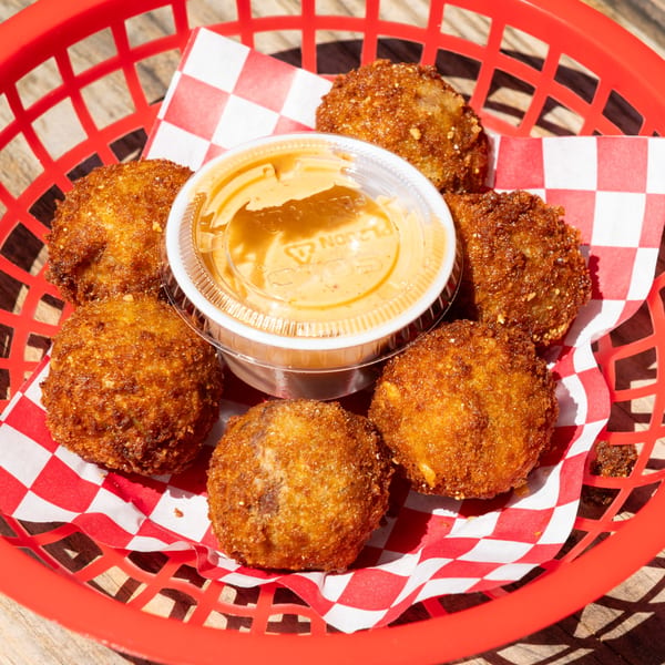 Six golden-brown fried hushpuppies and a small cup of dipping sauce in a red basket with checkered paper.