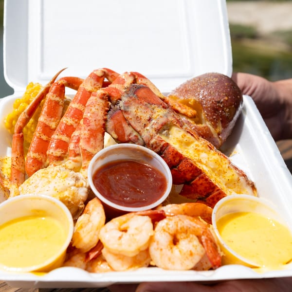 Seafood boil in a to-go container with crab legs, lobster, shrimp, corn, and dipping sauces.