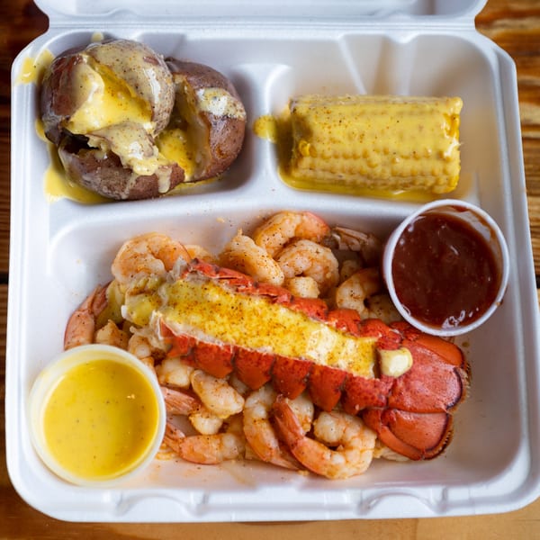 Seafood platter with lobster tail, shrimp, corn on the cob, and baked potatoes in a takeout container.