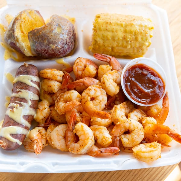 Shrimp boil with sausage, corn on the cob, and potato in a white takeout container.