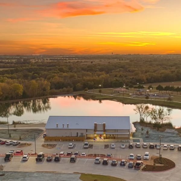 Aerial view of a restaurant building with a parking lot, lake, and park at sunset.