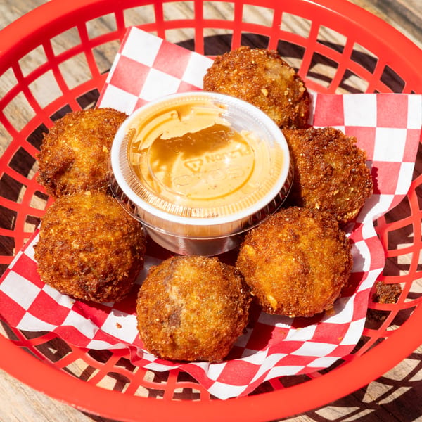 Six golden-brown fried hushpuppies arranged around a small cup of dipping sauce in a red basket.