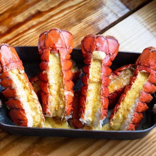 Four baked lobster tails with melted butter and seasoning in a black pan on a wooden table.