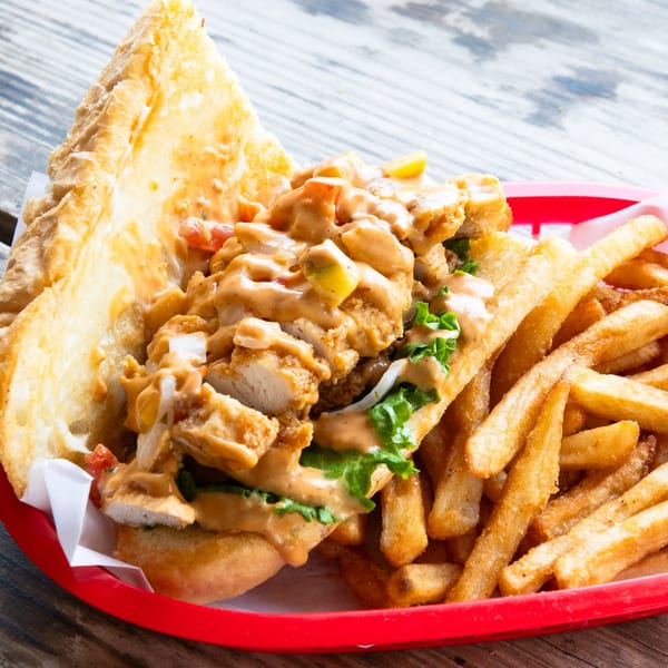 A fried chicken po'boy sandwich with lettuce, tomatoes, and sauce, served with fries.