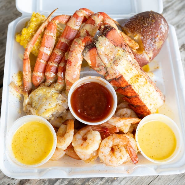 Seafood boil with crab legs, lobster, shrimp, corn, potato, and dipping sauces in a takeout container.