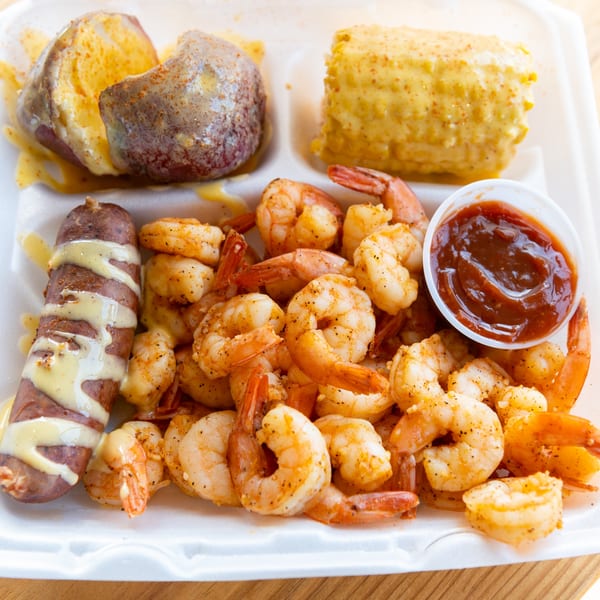 A takeout container filled with seasoned shrimp, a sausage link, baked potato, corn on the cob, and BBQ sauce.