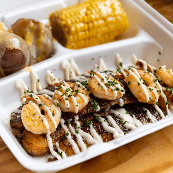 Shrimp and fish with creamy sauce, parsley, corn on the cob, and potatoes in a white takeout container.