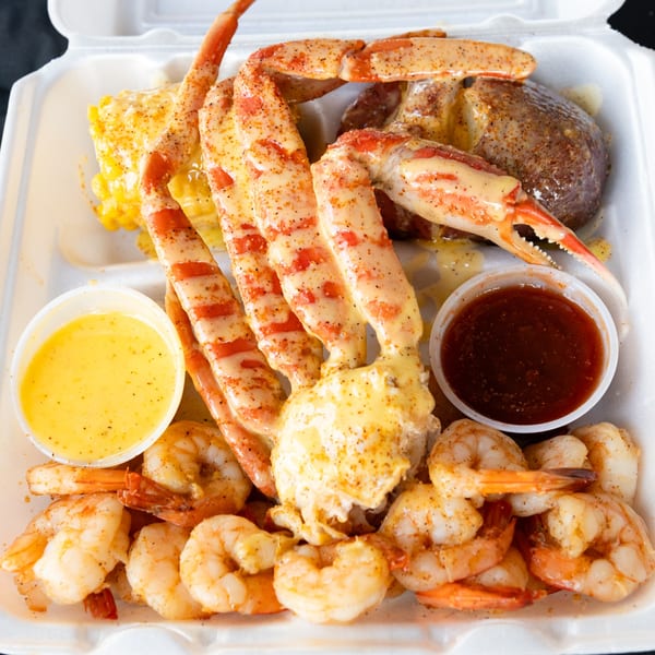 Snow crab legs, shrimp, corn, and potato in a take-out container with two dipping sauces.