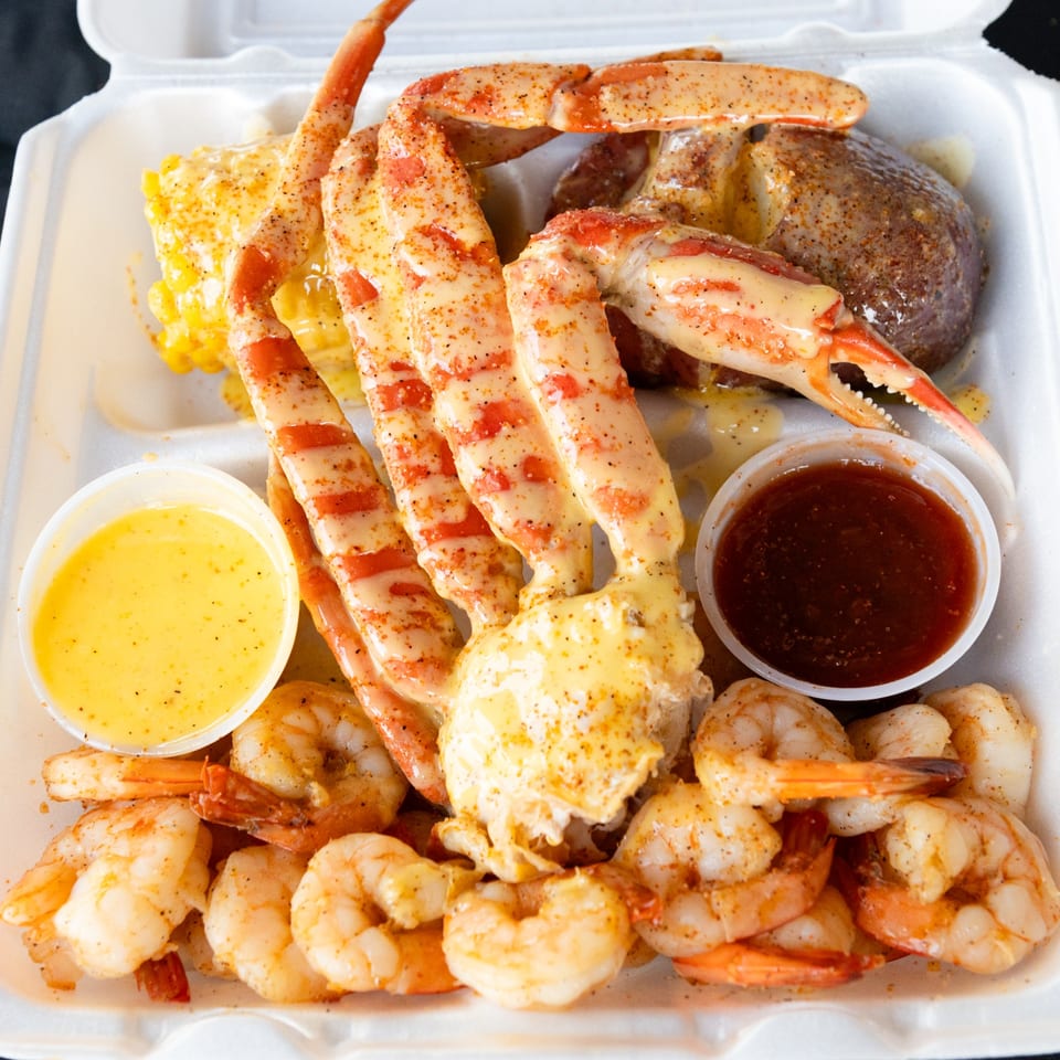 Snow crab legs, shrimp, corn, and potato in a take-out container with two dipping sauces.