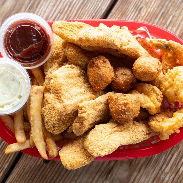 A red basket filled with fried fish, hush puppies, french fries, and two dipping sauces.