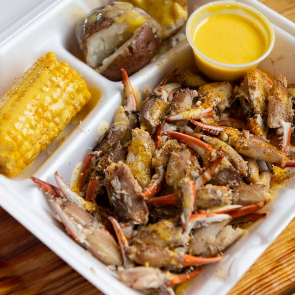 Crab legs, corn on the cob, and potatoes with sauce in a white takeout container.