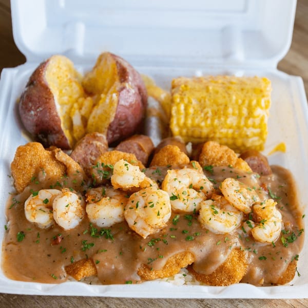 Shrimp and grits with gravy, corn on the cob, and red potatoes in a white takeout container.