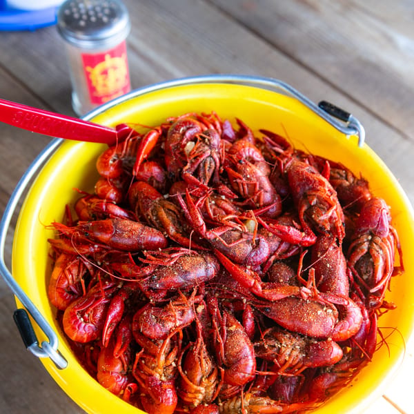A yellow bucket filled with seasoned, bright red boiled crawfish, with a red utensil and shaker nearby.