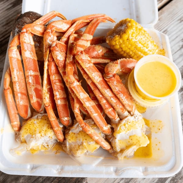 Snow crab legs, corn on the cob, and a side of butter sauce in a white takeout container.