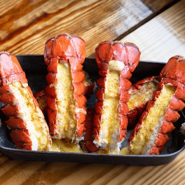 Four baked lobster tails with melted butter and seasoning in a black pan on a wooden table.