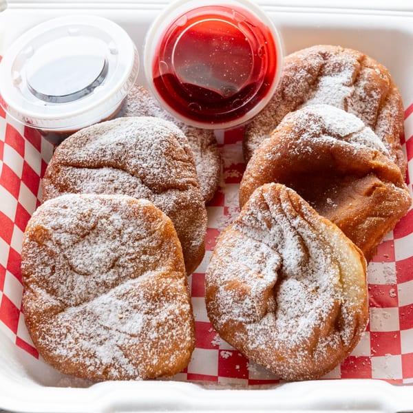Six powdered sugar beignets with raspberry and chocolate dipping sauces in a takeout container.