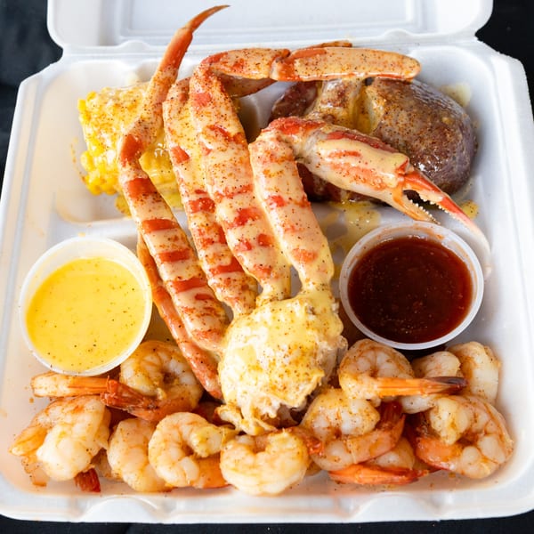 A seafood boil with crab legs, shrimp, corn, potato, and two dipping sauces in a takeout container.
