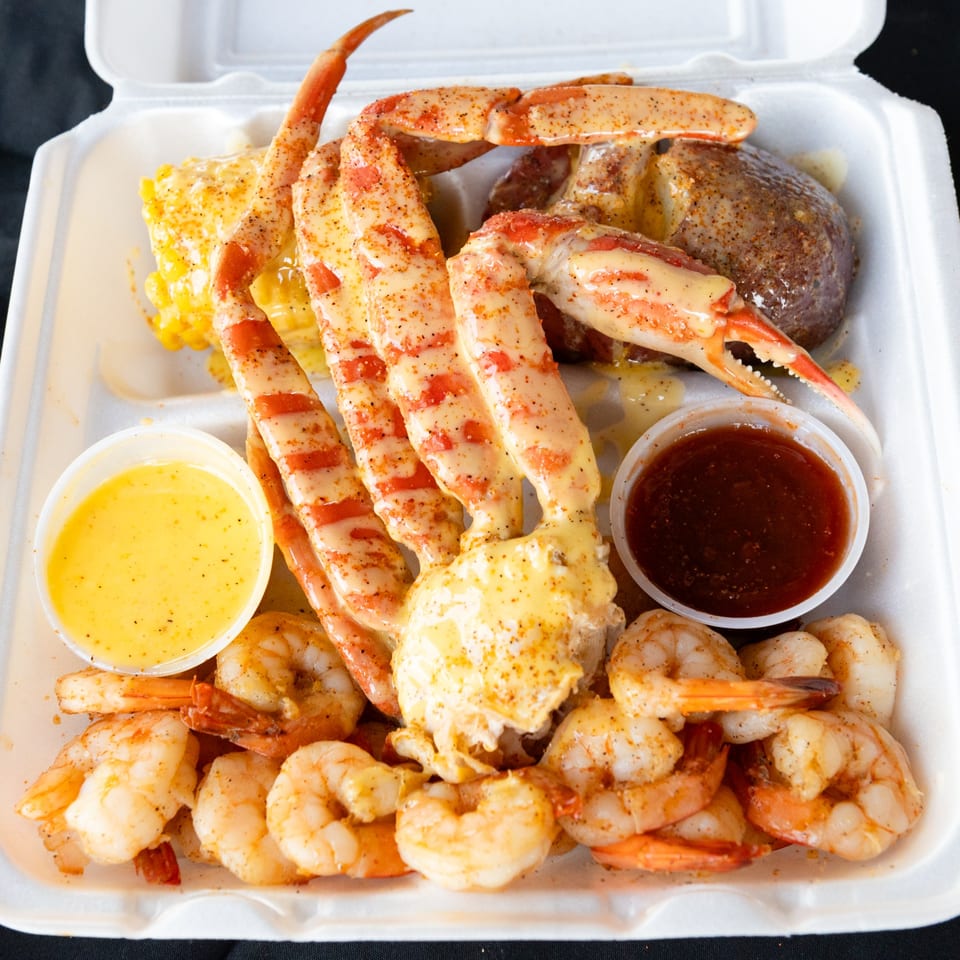 A seafood boil with crab legs, shrimp, corn, potato, and two dipping sauces in a takeout container.