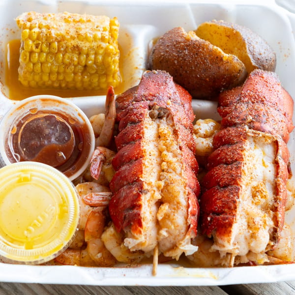 Seafood boil with two lobster tails, shrimp, corn on the cob, and a potato in a takeout container.