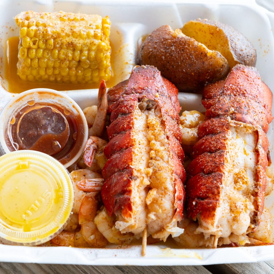 Seafood boil with two lobster tails, shrimp, corn on the cob, and a potato in a takeout container.