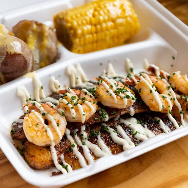 Shrimp and fish with creamy sauce, parsley, corn on the cob, and potatoes in a takeout container.