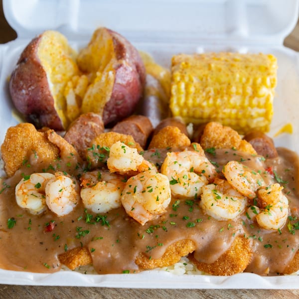 Shrimp and grits with fried fish, sausage, potatoes, and corn on the cob in a takeout container.