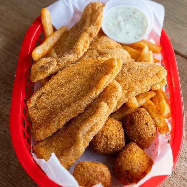Fried fish fillets, hush puppies, and french fries in a red basket with a side of tartar sauce.