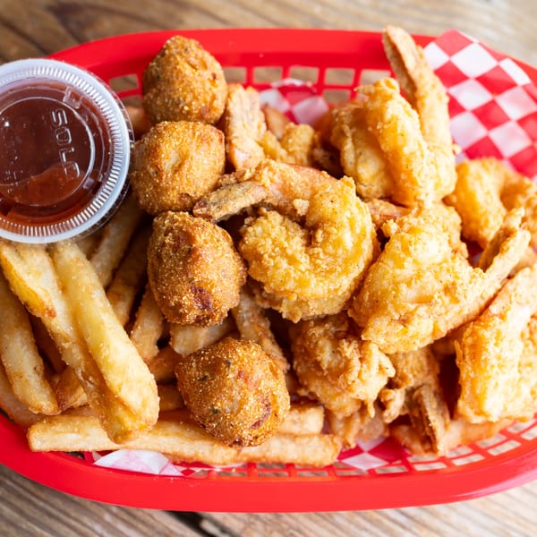Fried shrimp, hush puppies, and french fries in a red basket with a side of sauce.