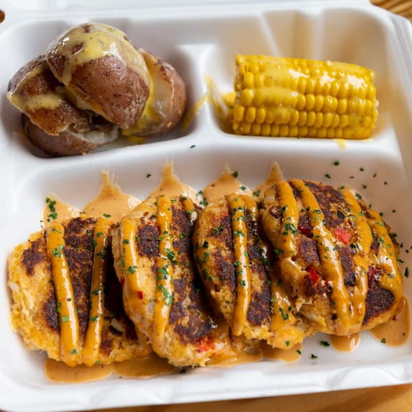 Crab cakes with sauce, roasted potatoes, and corn on the cob in a takeout container.