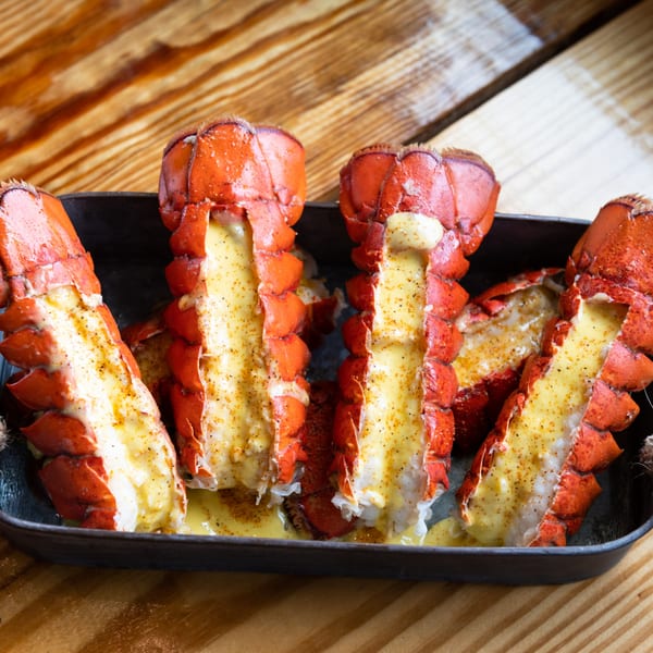 Four baked lobster tails with melted butter and seasoning in a black pan on a wooden table.